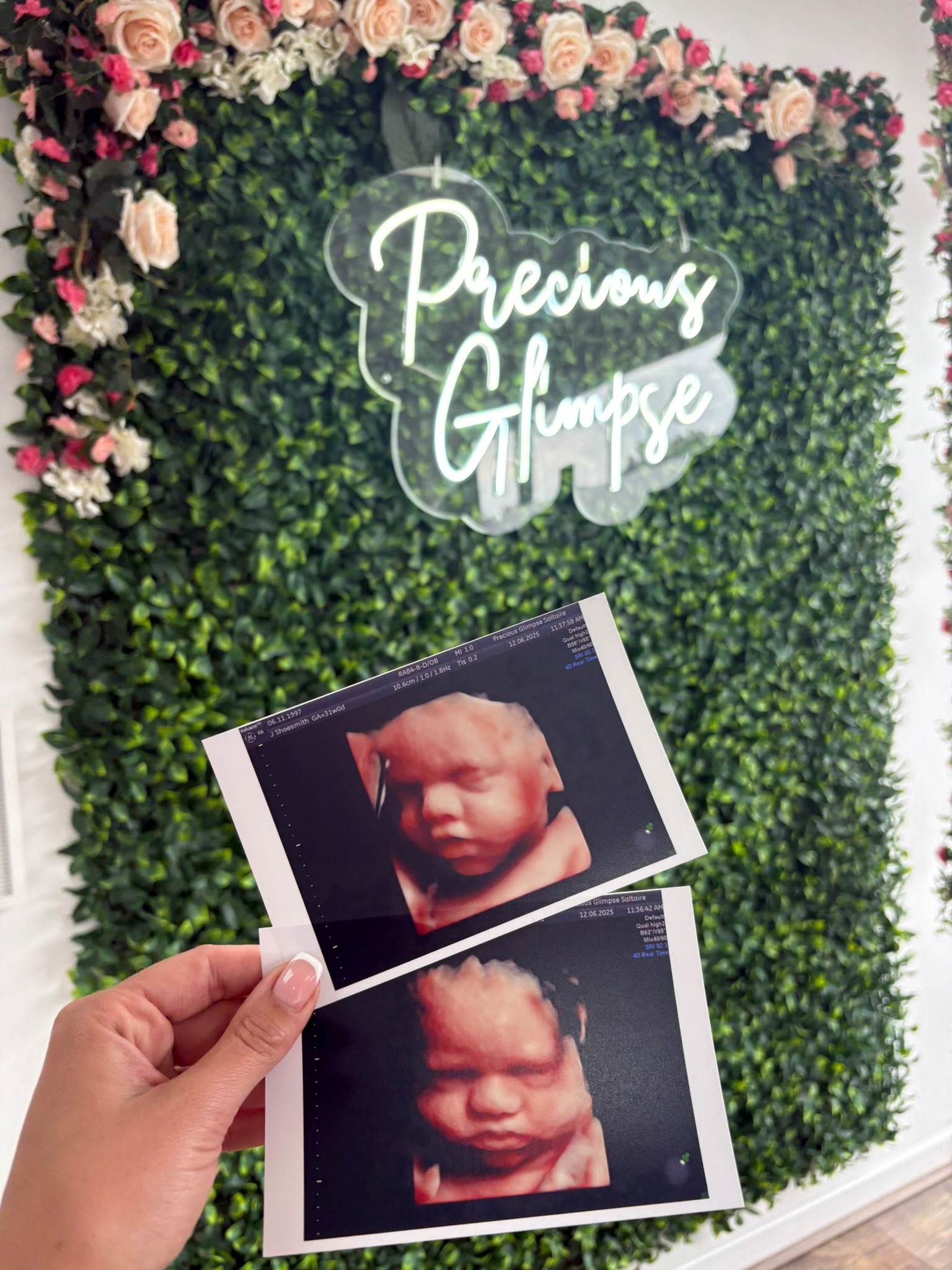 Hands holding 4D baby scan photos in front of the Precious Glimpse flower wall and neon sign