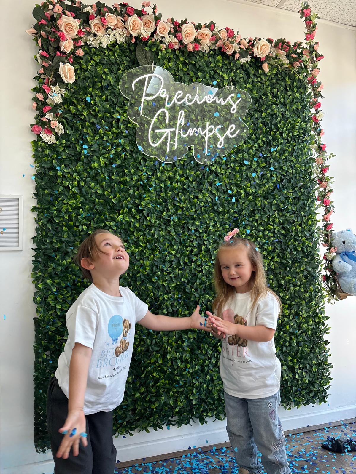 A brother and sister throwing blue confetti at the Precious Glimpse flower wall during a gender reveal