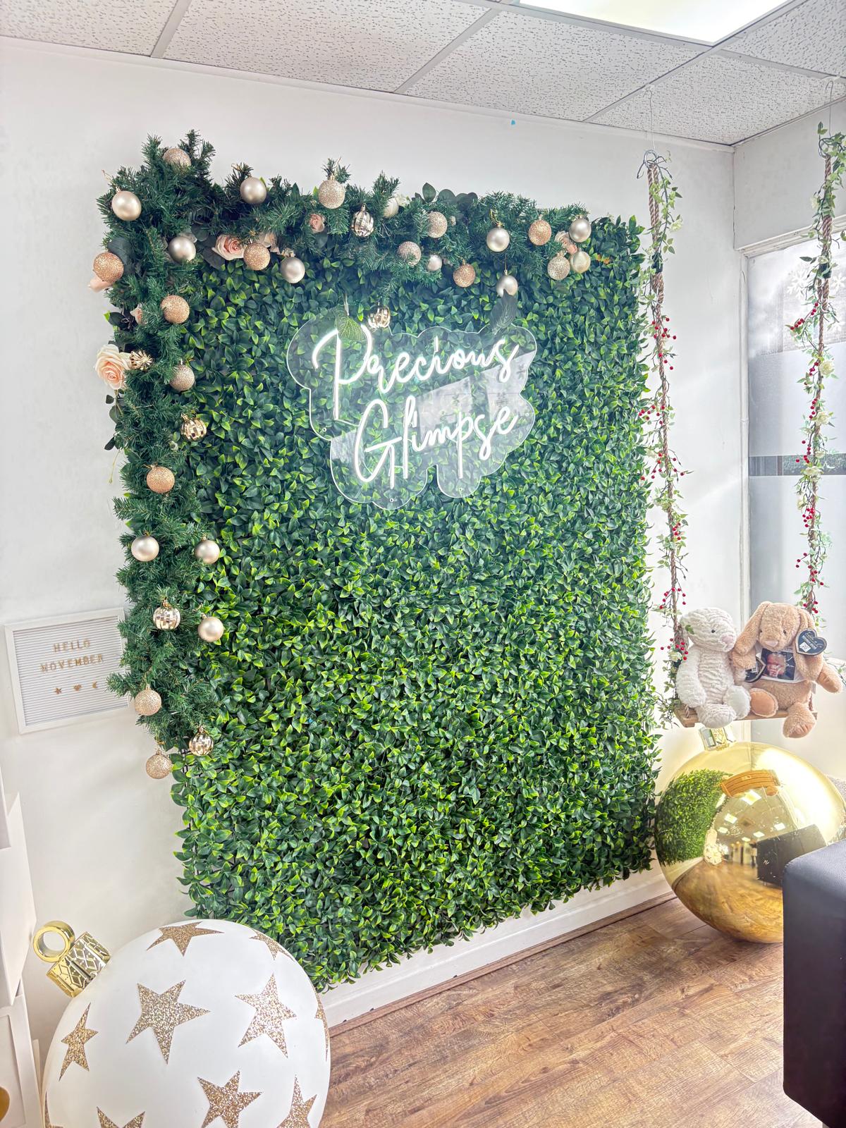 The Precious Glimpse neon sign on the studio's green flower wall, framed with festive garland and baubles